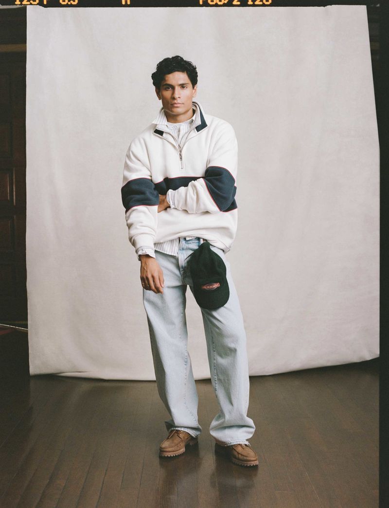 A young adult man poses in a photo studio wearing a white sweater, light blue jeans, and brown shoes, with a white backdrop.