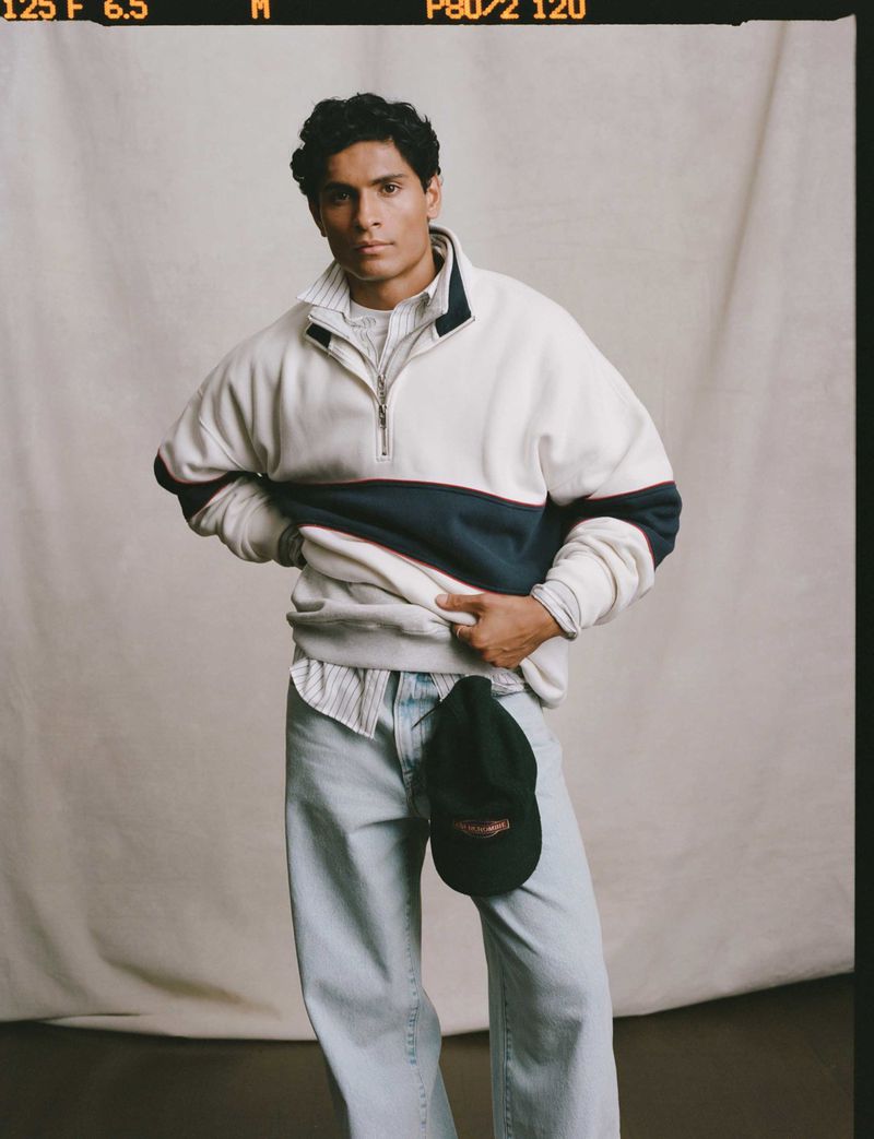 Young man in casual streetwear poses in front of a neutral backdrop. He's wearing a color-blocked sweatshirt, light wash jeans, and baseball cap.