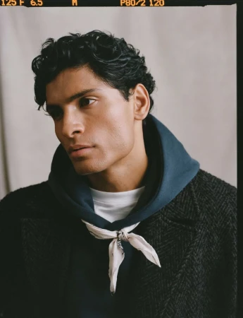 Adult male poses indoors wearing a hooded sweatshirt, patterned bandana, and herringbone coat in a medium shot with neutral lighting.