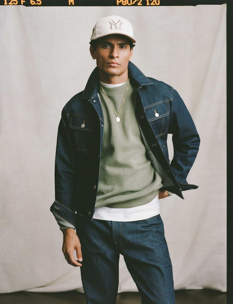 Full-length portrait of a young adult man in a denim jacket, sweatshirt, jeans, and baseball cap against a neutral backdrop.