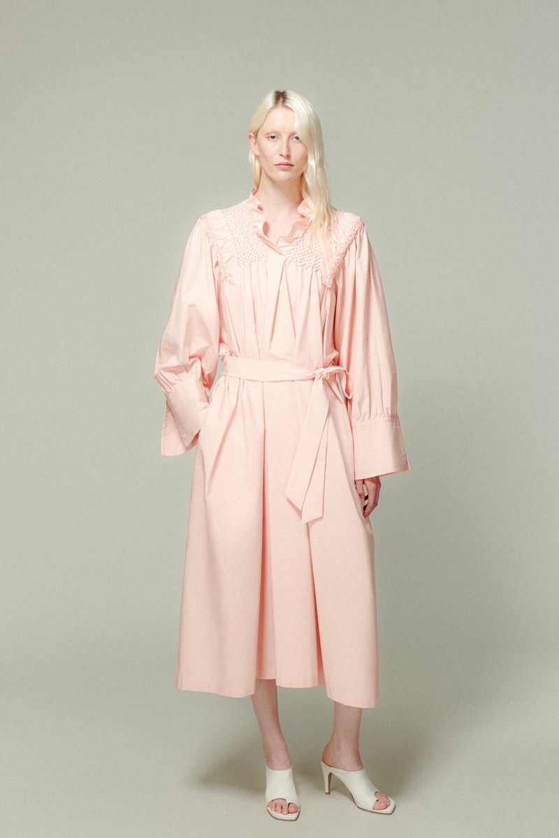 Full-length shot of a light-skinned young adult female model with platinum blonde hair wearing a pink dress and white heeled sandals.