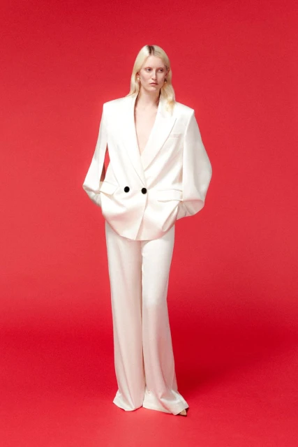 A fair-skinned woman with long blonde hair poses in a white blazer and slacks against a bright red backdrop in a fashion editorial.