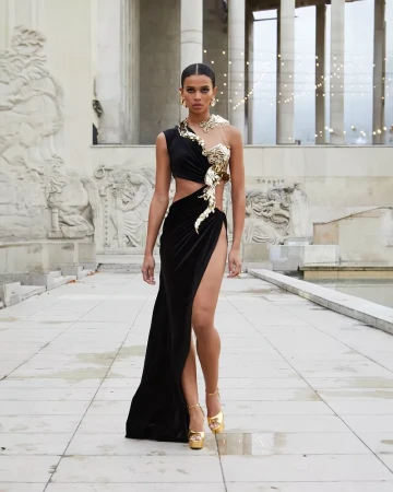 Elegant woman in a black and gold dress, posing outdoors in front of a building with sculptures. Fashion, beauty, and architecture.