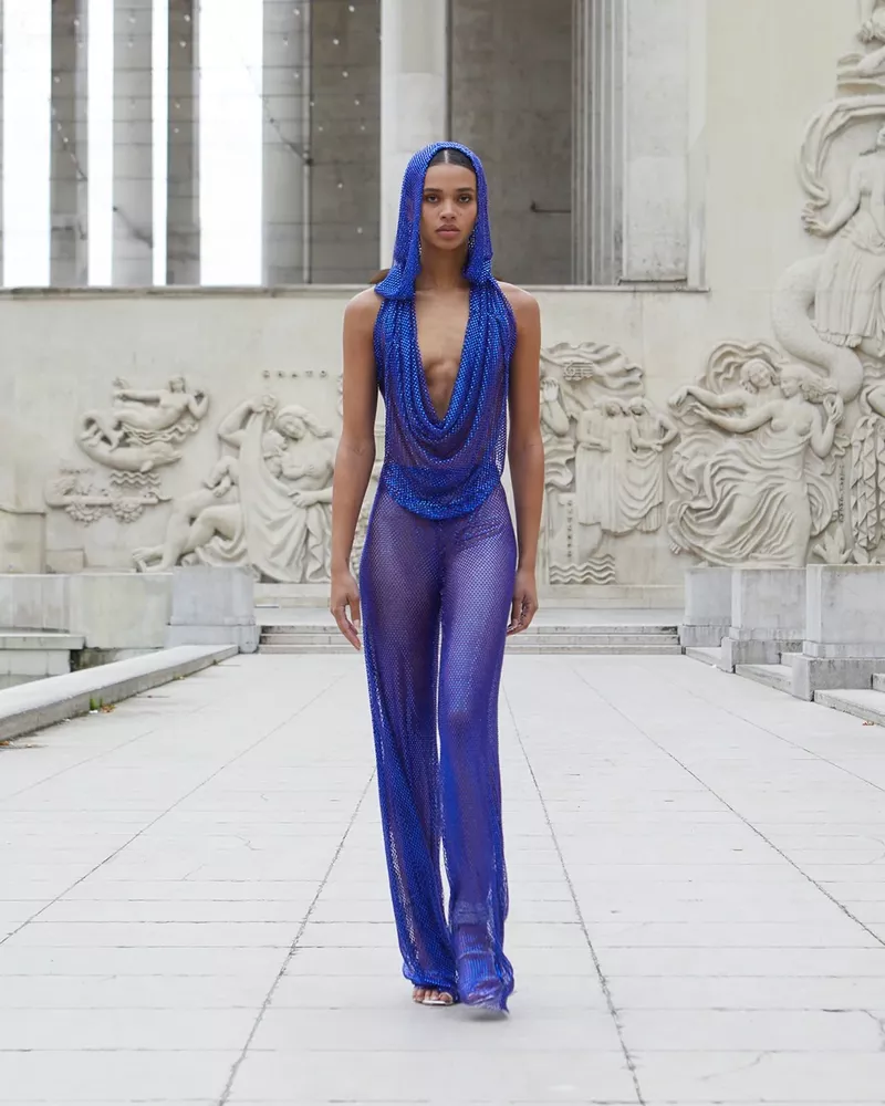 A woman walks on a tiled path wearing a blue sequin hooded top and pants, set against an outdoor backdrop of carved stone reliefs.