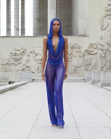 A woman walks on a tiled path wearing a blue sequin hooded top and pants, set against an outdoor backdrop of carved stone reliefs.