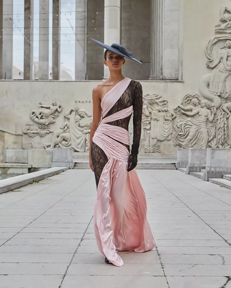 A runway model in a one-shoulder pink gown with lace panels poses in front of an ornate building for a fashion editorial photograph.