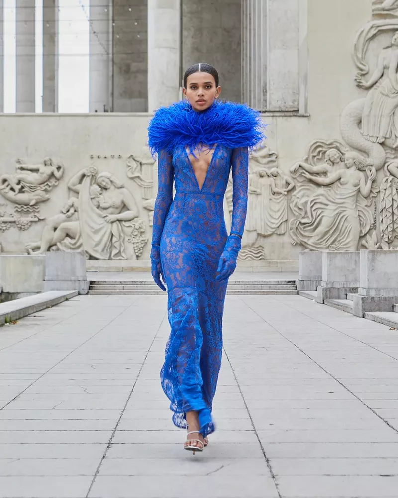 Woman in elegant blue lace gown with feathered collar walks a stone path near a building with relief sculpture.