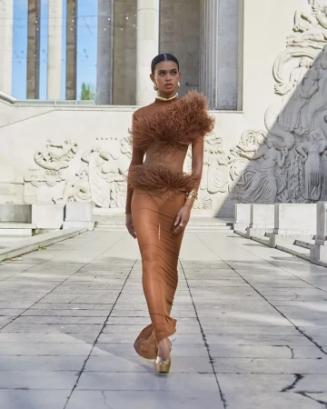 A woman in a brown, feathered, sheer dress walks a light stone path with neoclassical architecture in the background on a sunny day.