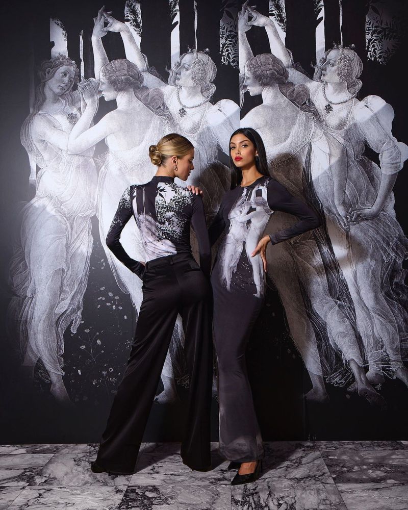 Two fashion models pose in elegant long-sleeved dresses with dark graphics against a matching black and white painted backdrop in a studio.