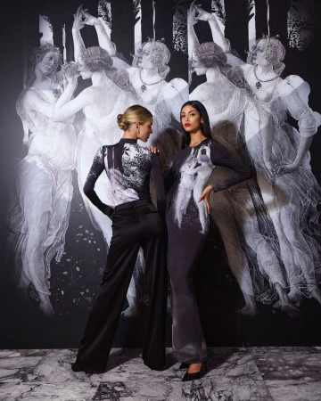 Two fashion models pose in elegant long-sleeved dresses with dark graphics against a matching black and white painted backdrop in a studio.