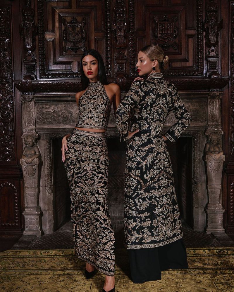 Two women in ornate black and gold gowns pose in an elegant, dark interior with a carved wooden wall and fireplace, fashion photography.