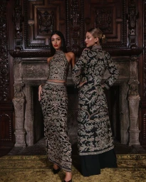 Two female models in patterned black and gold two-piece outfits posing before an ornate carved wooden fireplace.
