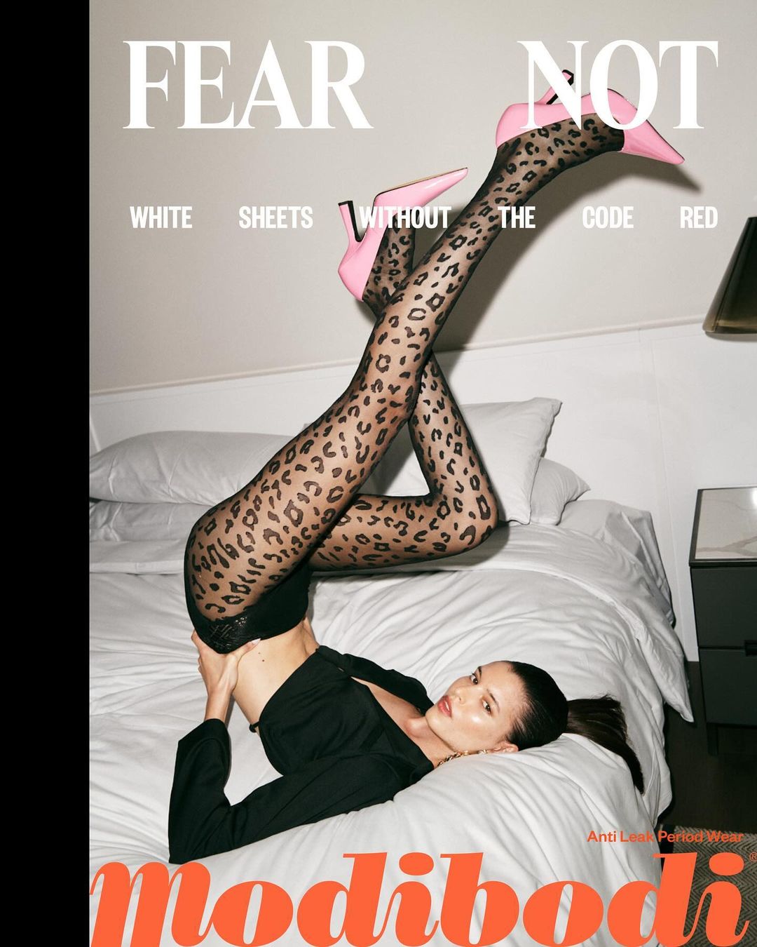 A woman lies on a bed with white sheets, wearing patterned hosiery and pink heels, posing for a fashion advertisement campaign.