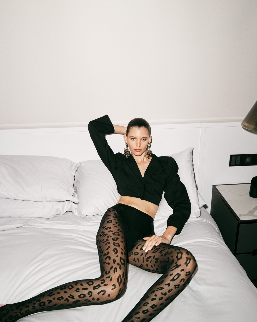 A woman reclines on a bed wearing a black cropped jacket and patterned tights, illuminated by studio lighting, in a fashion-forward pose.