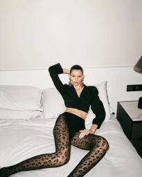 Model wearing black crop top and animal print tights seated on a white bed in a hotel room interior.