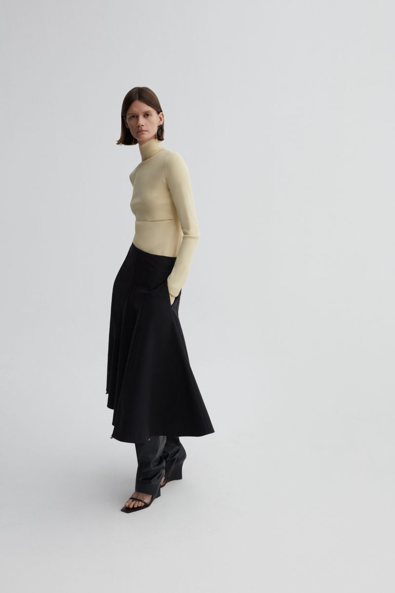 A woman poses in a studio wearing a yellow long-sleeve shirt, black skirt, and black wedge sandals against a white backdrop.