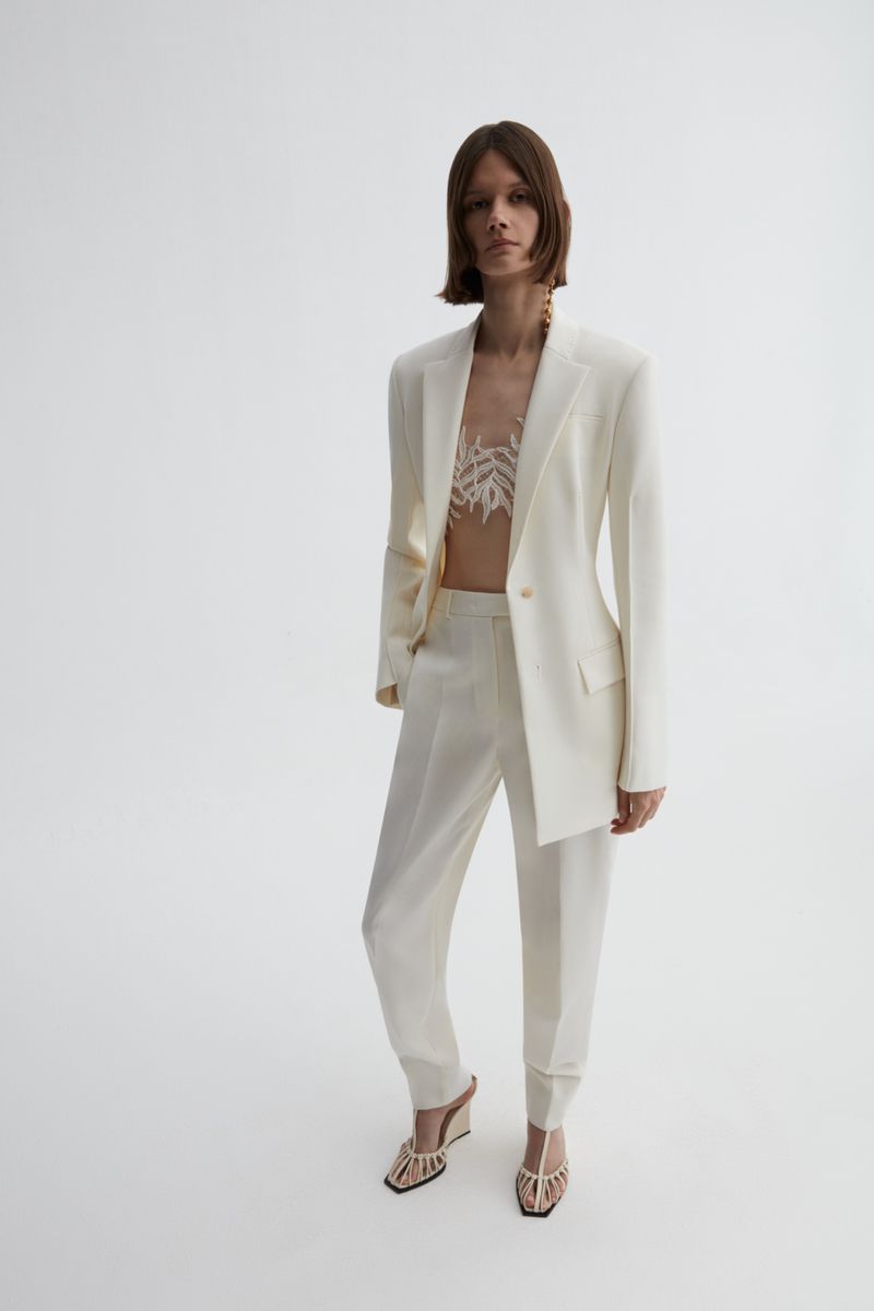 Full-length studio shot of a woman in an off-white suit with decorative top and sandals against a neutral background.