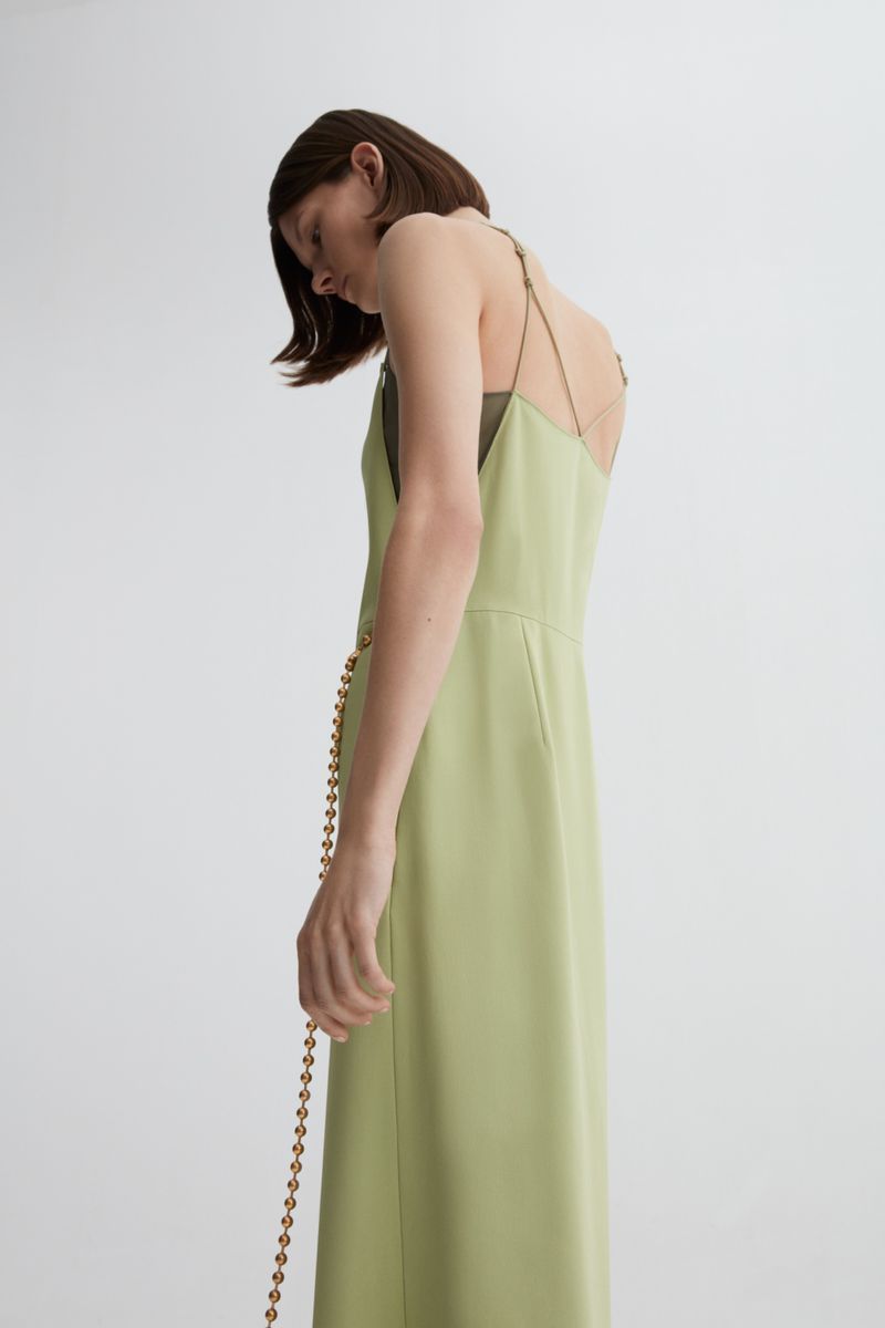 Adult woman wearing light-green dress with spaghetti straps, posing in a neutral, indoor setting. Fashion editorial shot.
