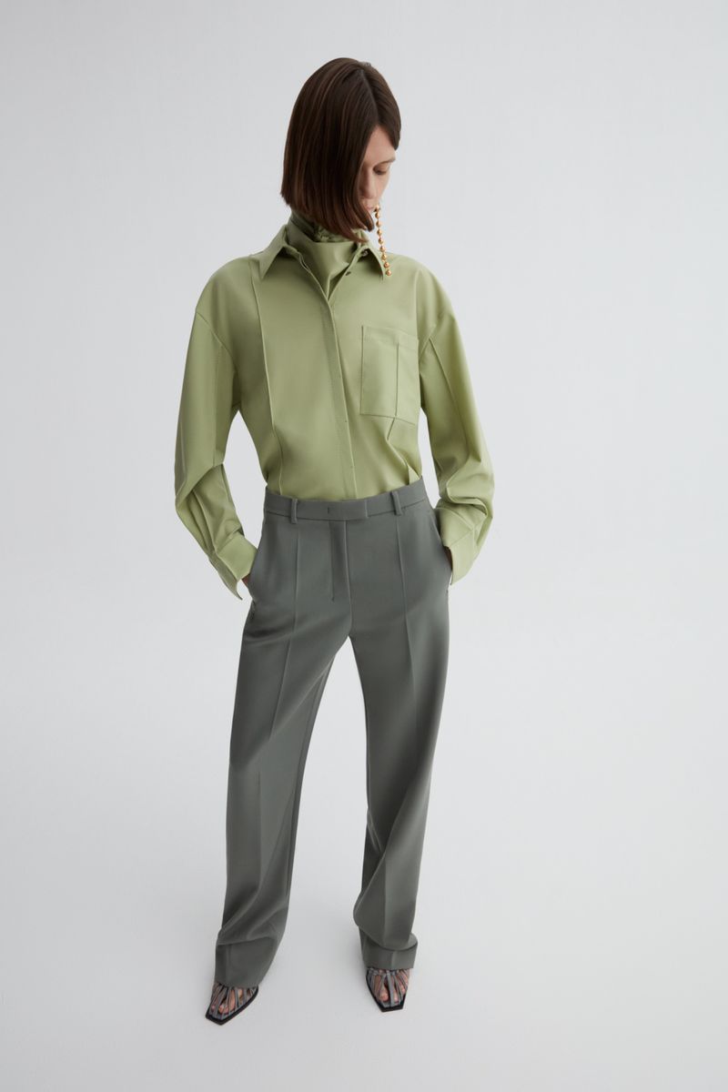 Model wearing a sage green button-down shirt and grey trousers against a white seamless studio backdrop.