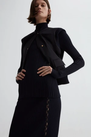 Fashion shot of a woman wearing a black turtleneck sweater, skirt, and vest in a studio with a neutral background in portrait orientation.