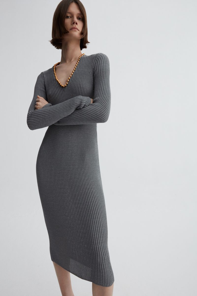Adult woman in a gray ribbed, long-sleeve midi dress with a gold beaded necklace poses indoors against a neutral background in a studio shot.