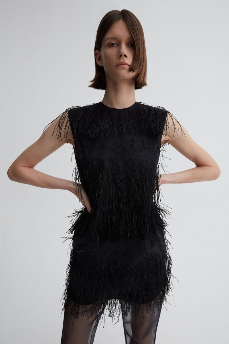 A woman poses in a black sleeveless dress with fringe detail against a white backdrop in a studio setting; full studio lighting.