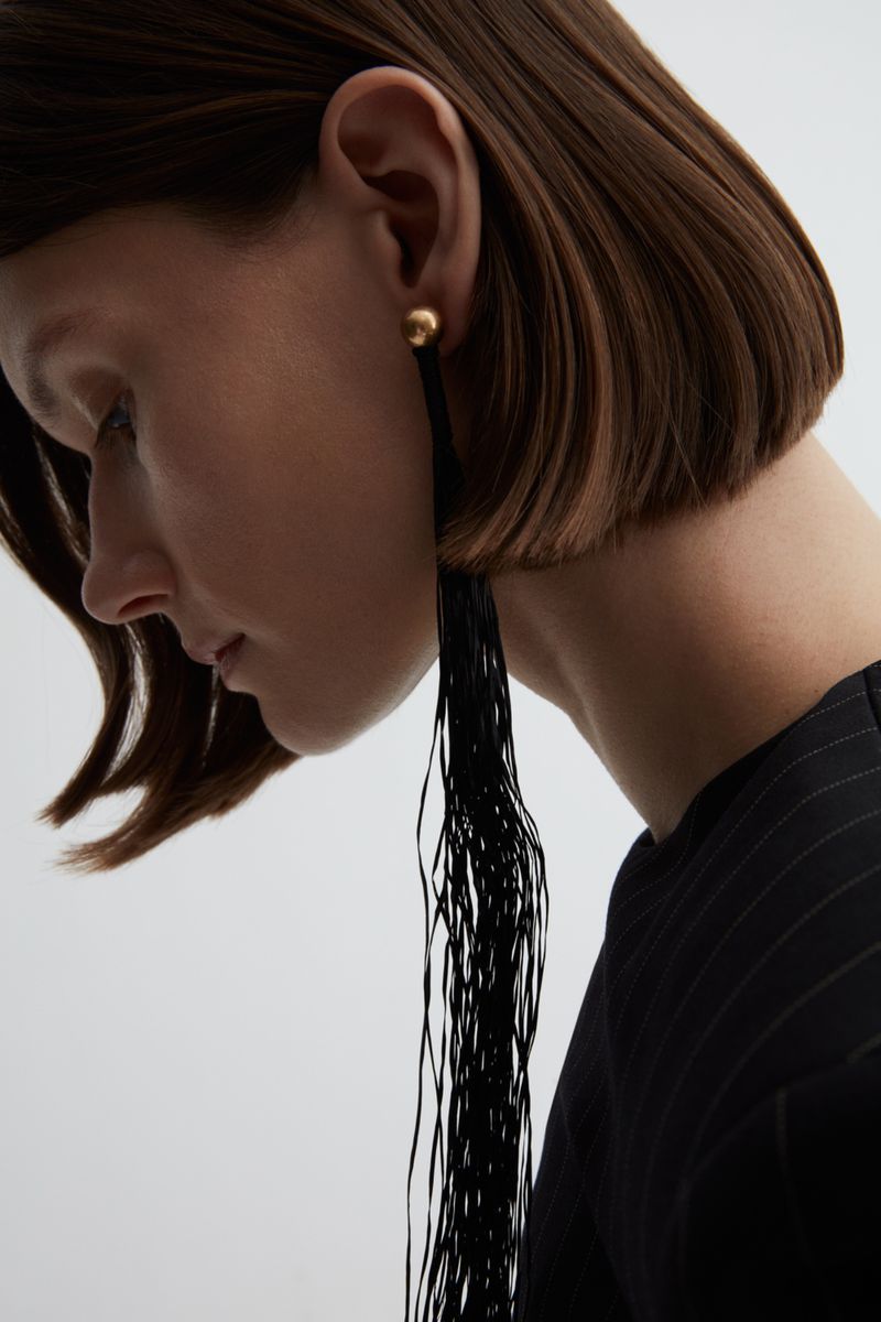 Close-up of a woman with short brunette hair and fair skin, wearing a black top, and dangling black and gold earrings, in a studio shot.
