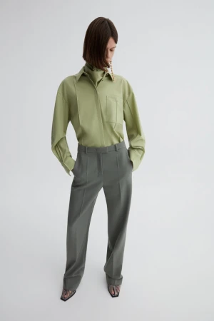 Model wearing a sage green button-down shirt and grey trousers against a white seamless studio backdrop.