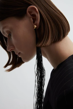 Close-up profile of a woman wearing a long black fringe earring and a dark pinstripe top against a white backdrop.