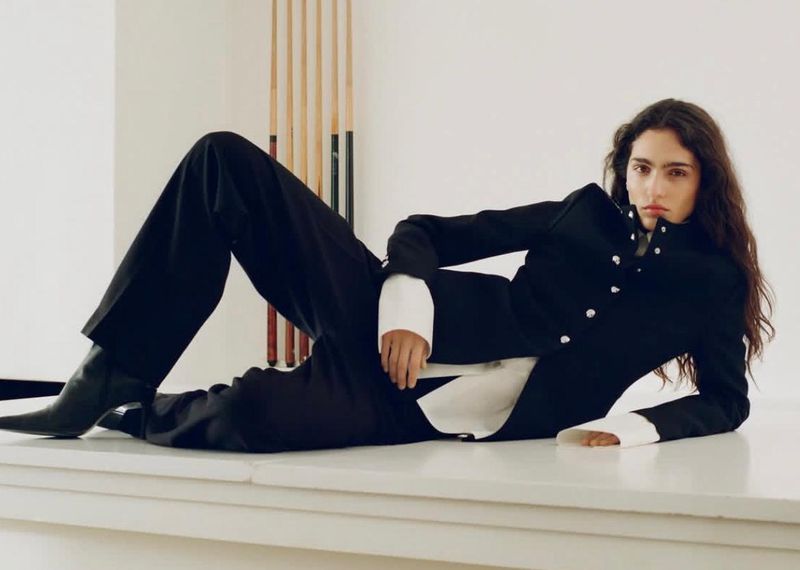 A brunette fashion model in a dark suit and black boots reclines on a white surface in a minimalist studio with pool cues in the background.