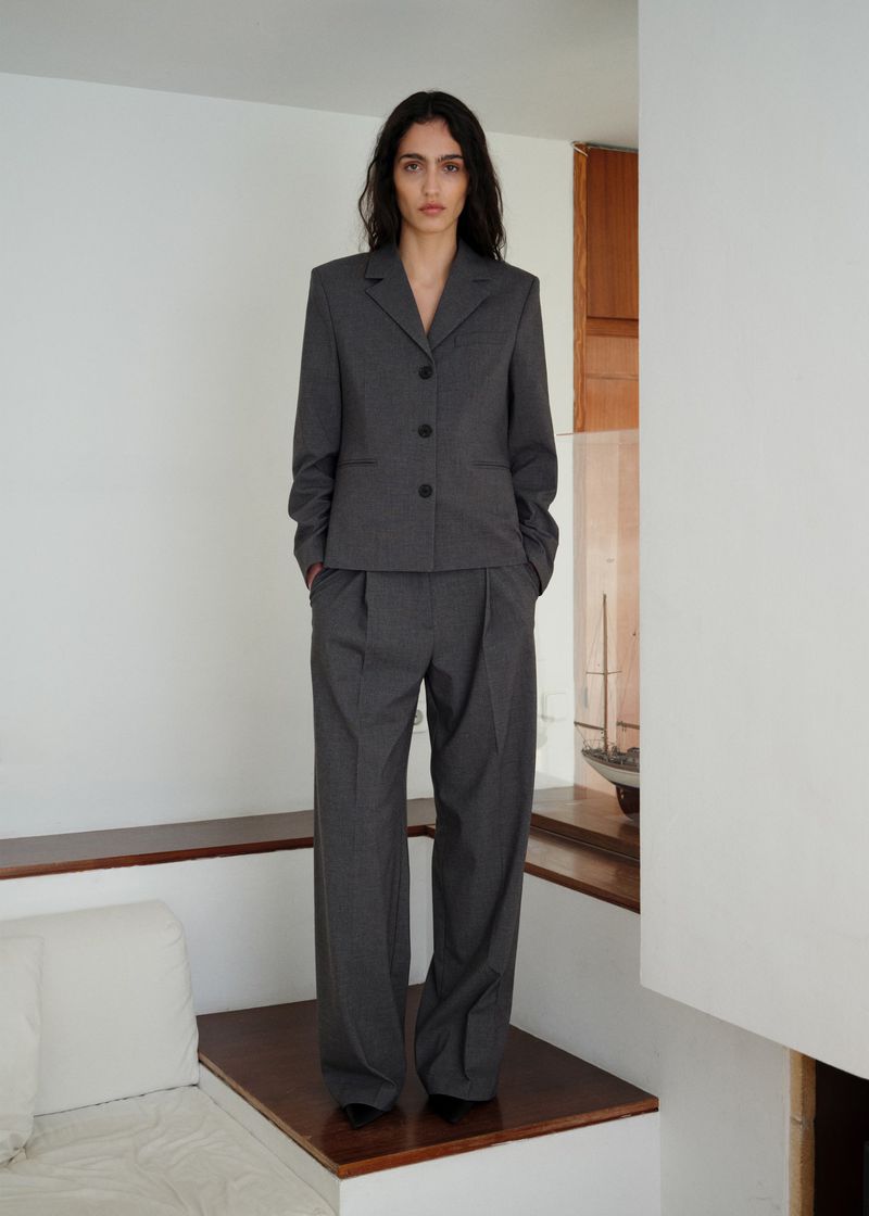 Model wearing a grey tailored suit jacket and matching trousers standing indoors in a minimalist interior space.