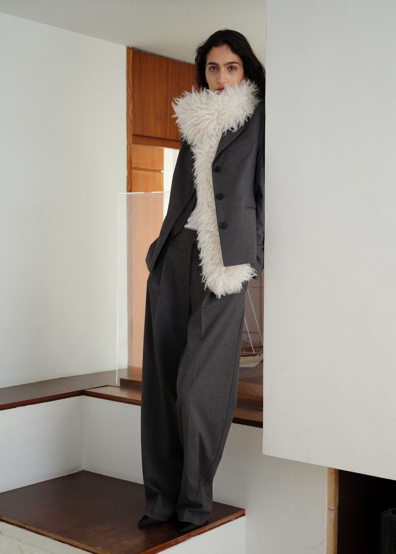 A woman poses indoors in a grey suit with a white, furry stole. The setting features white walls and minimalist architecture.