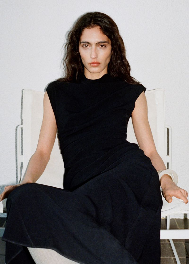 Model wearing a black sleeveless dress seated on a white chair against a plain white wall