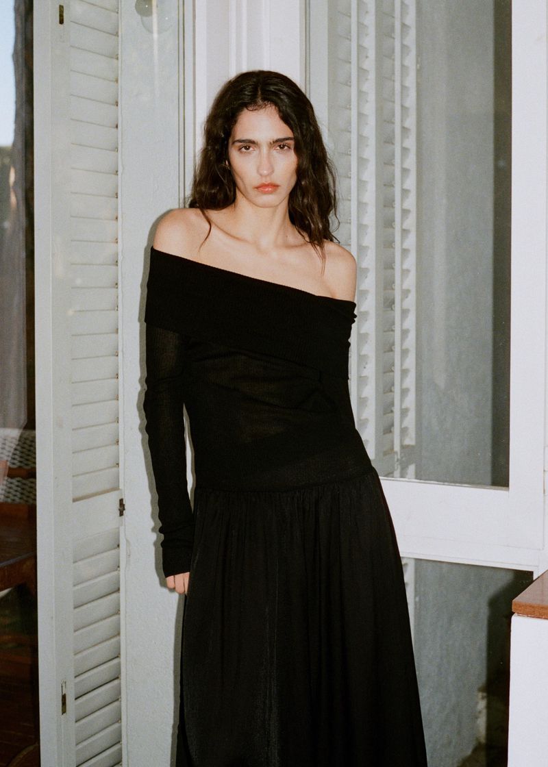 Fashion portrait of a woman in a long-sleeve black off-shoulder top and long black skirt standing against a white shuttered window.