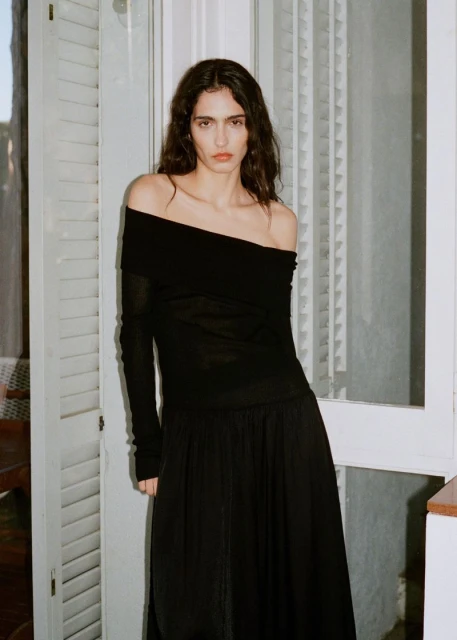 Fashion portrait of a woman in a long-sleeve black off-shoulder top and long black skirt standing against a white shuttered window.