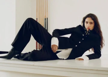 Model lying on a white surface wearing a black jacket and trousers with white cuffs, against a plain background.