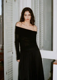 Model wearing an off-the-shoulder black dress standing indoors near white louvered doors for Mango AW 25.