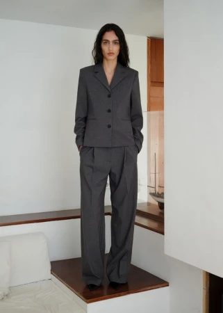 Model wearing a grey tailored suit jacket and matching trousers standing indoors in a minimalist interior space.