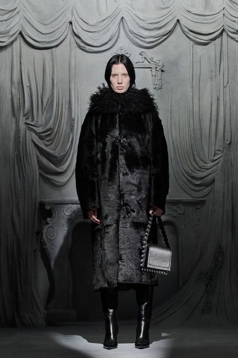 A woman with dark hair poses indoors wearing a long black coat with a fur collar, black boots, and holding a black bag against a gray backdrop.