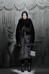 Model wearing a black fur coat and holding a black handbag in front of draped grey set-dressed background