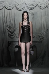 Model in a black strapless cutout mini dress holding a handbag against a draped classical sculpted wall backdrop