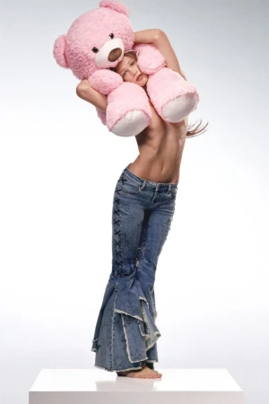 Adult woman embraces large pink teddy bear while standing on white cube, wearing flared jeans against white backdrop in a studio shot.