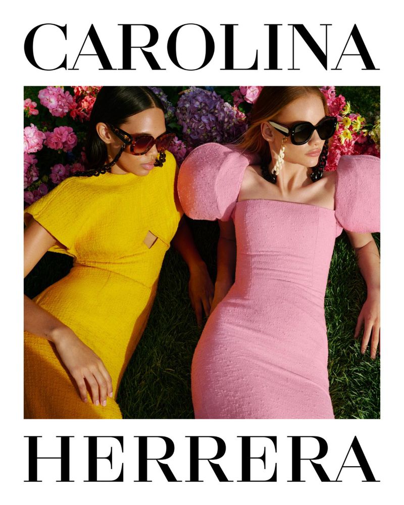 Two fashion models in pink and yellow dresses pose on green grass among colorful flowers, wearing sunglasses for a Carolina Herrera ad.