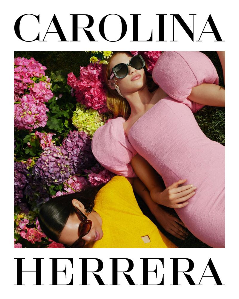 Two women in brightly colored dresses are lying in a field of colorful flowers. The Carolina Herrera logo is prominently displayed.
