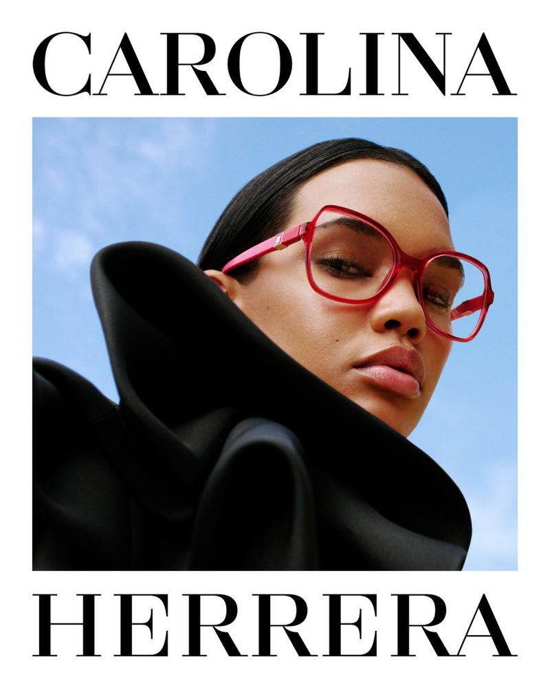 A close-up of a woman wearing pink glasses and a black ruffled collar, set against a blue sky, framed by 'Carolina Herrera' text.