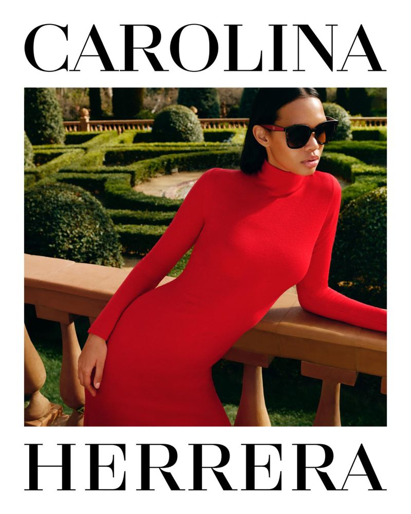 Woman in a red dress and sunglasses poses outdoors against a garden backdrop for Carolina Herrera fashion brand promotion.
