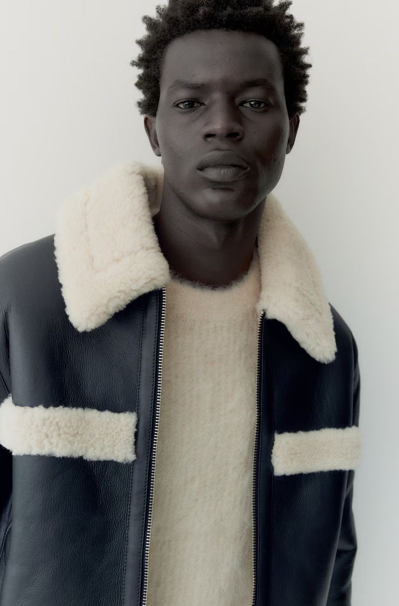 A dark-skinned male model in a beige sweater and black shearling jacket poses in a studio with a neutral background.