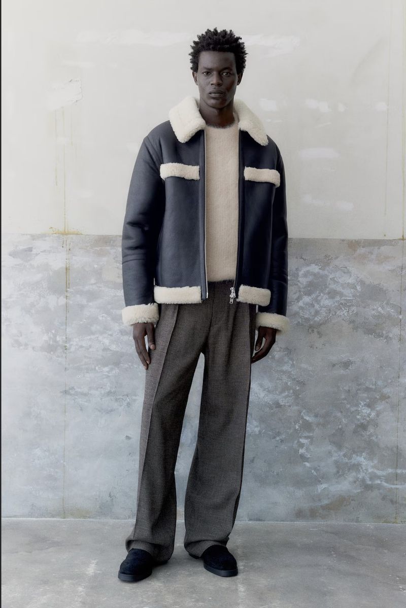 A stylish Black male model poses indoors wearing a navy shearling jacket, beige sweater, and wide-leg grey pants for a fashion editorial.