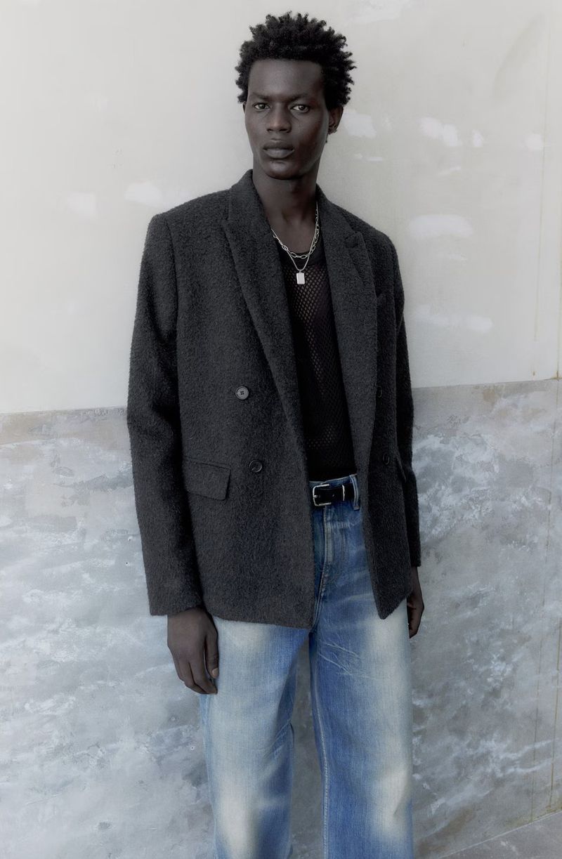 Black man wearing dark grey blazer, black mesh top, and blue jeans against a textured wall