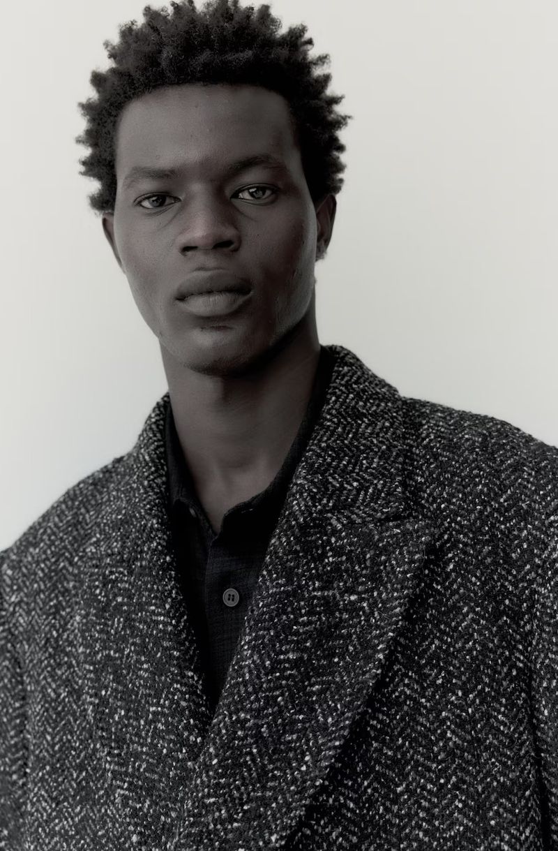 Portrait of an adult male with dark skin wearing a herringbone overcoat in front of a plain light grey backdrop in a studio setting.
